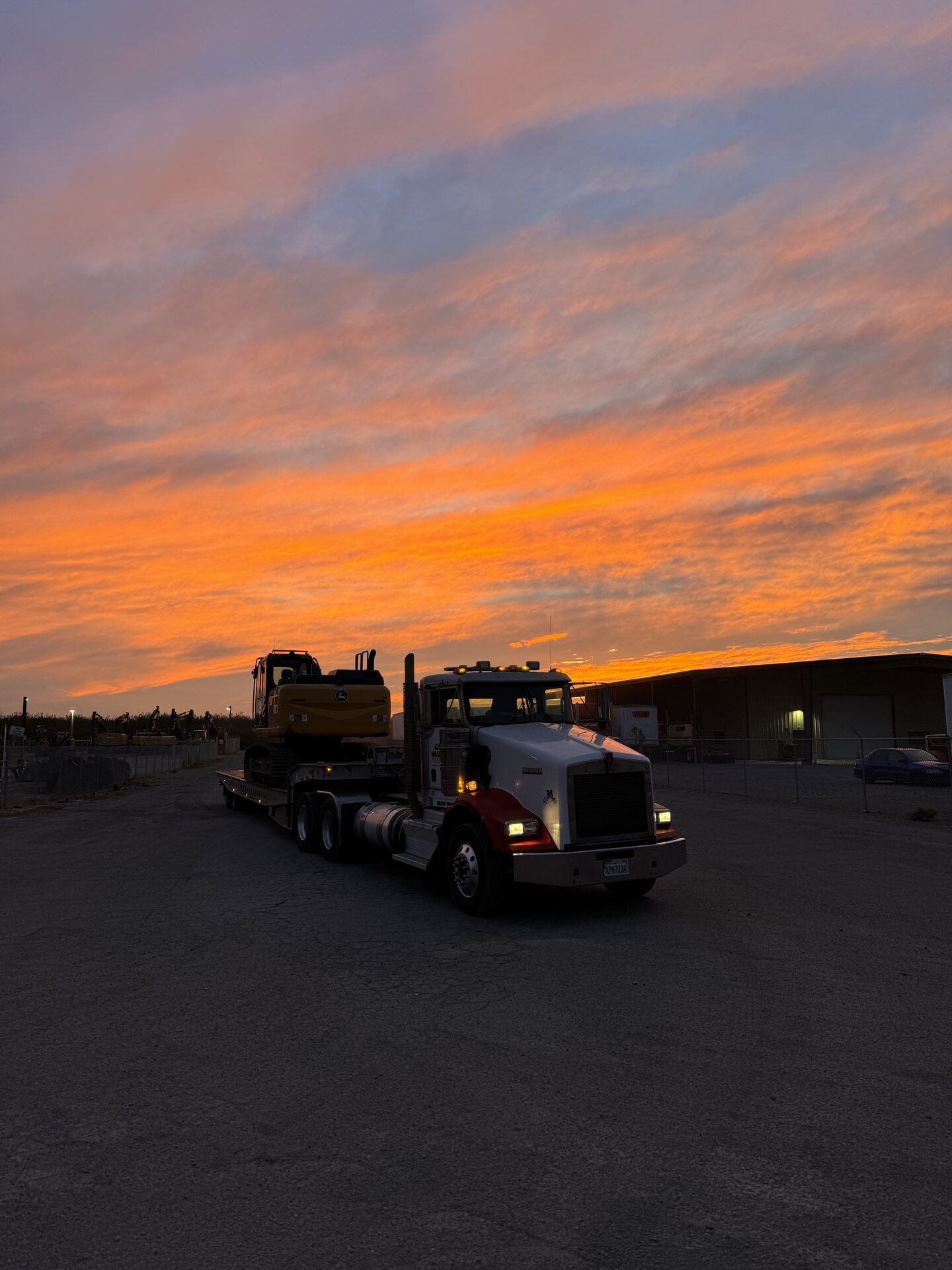 CKJ Ag Services Kenworth flatbed hauling a John Deere excavator at sunset